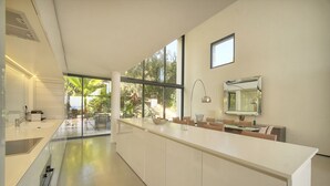 Una cocina moderna con una isla grande, un comedor con sillas y ventanas amplias que ofrecen vista a vegetación.