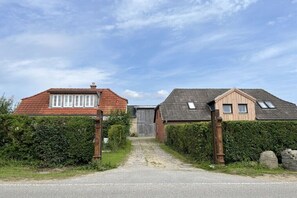 Eine ländliche Straße führt zu einem Anwesen mit einem Haus mit rotem Dach und einem Gebäude mit grauem Dach, umgeben von Hecken.