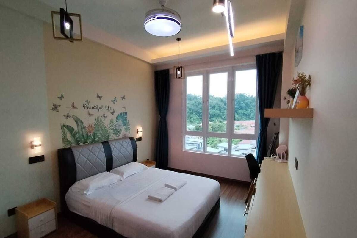 A bedroom with a bed, bedside tables, a nightstand, a window with curtains, and a view of greenery outside.