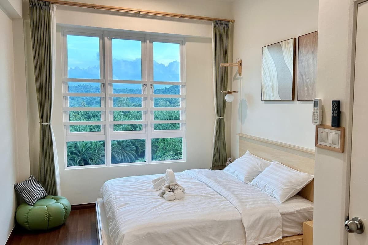 A bedroom with a large window, a bed, a bedside table, a lamp, and a door.