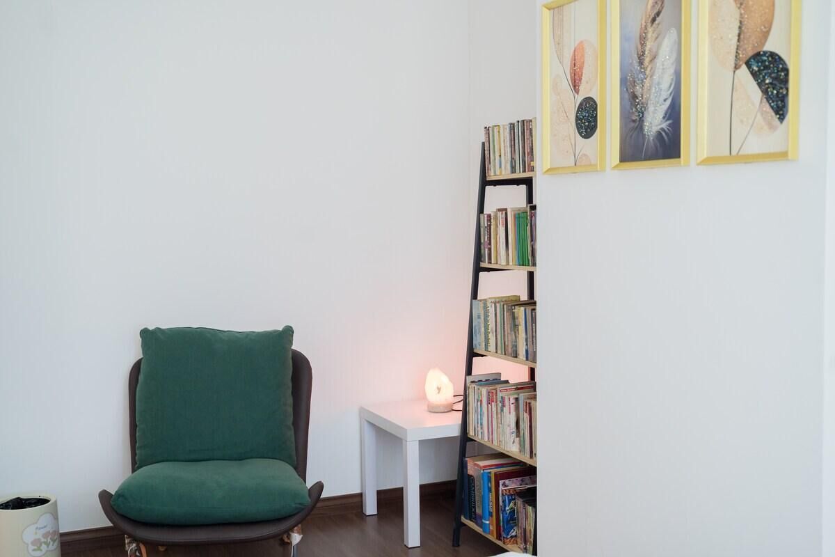 A room with a green chair, a white table with a lamp, and a bookshelf with books.