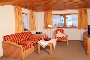 Ein gemütliches Wohnzimmer mit einer Couch, einem Sessel, einem Tisch mit Lampe und einem Fenster mit Vorhängen.