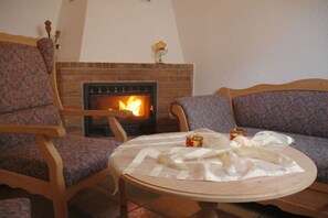 A cozy living room with a fireplace, two patterned chairs, a round table with a white cloth, and a vase with flowers. - Holiday home in Briedern with swimming pool (Briedern)