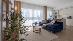 Un salon moderne avec un canapé bleu, deux tables rondes et une grande fenêtre ornée de rideaux légers.