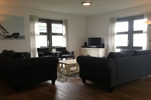 A living room with a sofa, chairs, a coffee table, a television, and large windows. - Villa 78 in Villapark Sneekermeer (Offingawier)