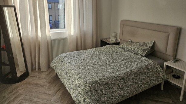 Ein Schlafzimmer mit einem Bett, einem Nachttisch, einem Spiegel und einem Fenster mit Vorhängen.