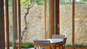 A modern interior with a sofa, dining table, and chairs, a stone wall, and a view of greenery outside.