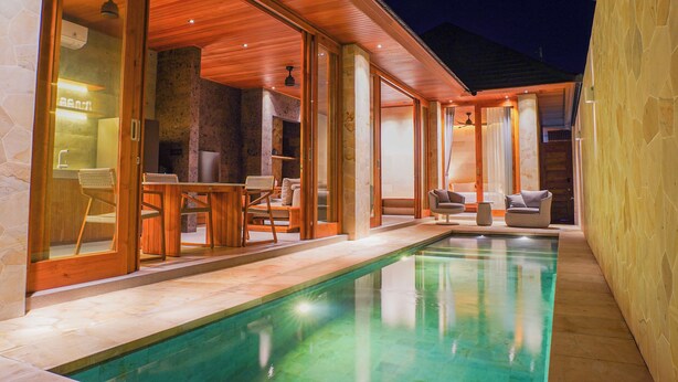 A swimming pool with a wooden deck, glass doors leading to a seating area with chairs and a table, and a stone wall on one side.
