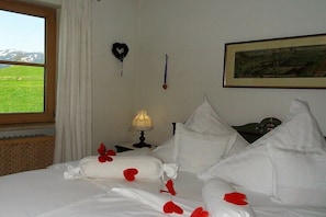 Ein Hotelzimmer mit Bett, Nachttisch mit Lampe, einem gerahmten Bild an der Wand und einem Fenster mit Blick auf ein grünes Feld.