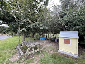 A picnic area with a wooden table and benches, surrounded by trees and a small yellow shed.