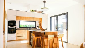 Eine moderne Küche mit Holzausstattung, einem Einbauofen, einem großen Fenster und einer Essecke mit Holzstühlen.