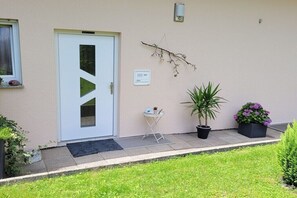 Una entrada de edificio con una puerta blanca, una mesa pequeña con una planta, y una maceta con flores moradas.