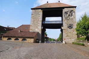 Ein steinernes Tor mit einem Balkon, einem Wappen und einem Gebäude mit einem Ziegeldach im Hintergrund.