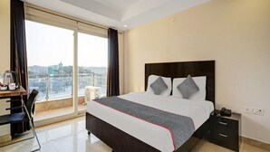 Quarto de hotel com cama, escrivaninha, cadeira, mesa de cabeceira e vista para a cidade.