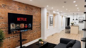 Ein modernes Wohnzimmer mit Backsteinwand, Flachbildfernseher, einer Couch, einem Couchtisch und einer Küchenzeile im Hintergrund.