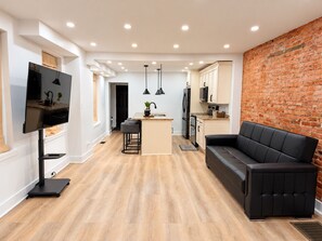 Ein modernes Wohnzimmer mit einer schwarzen Ledersofa, einem Flachbildfernseher an einer Halterung und einer Küchenzeile mit Backsteinwand.