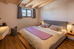Ein Schlafzimmer mit einem großen Bett, einem Fenster mit Vorhängen, einem Nachttisch mit Lampe und einem kleinen Hocker.