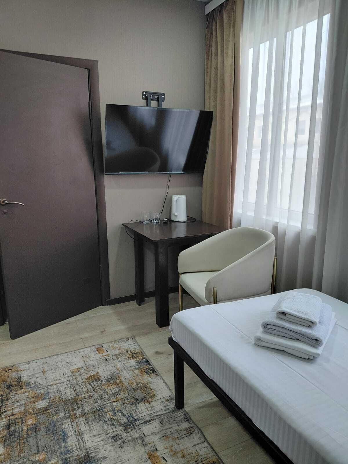 A hotel room with a bed, a TV mounted on the wall, a small table with a kettle, a chair, and a door.
