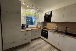 Eine moderne Küche mit hellen Holzschränken, einem eingebauten Ofen, einem schwarzen Spülbecken und einem Fenster mit Blick auf Bäume.
