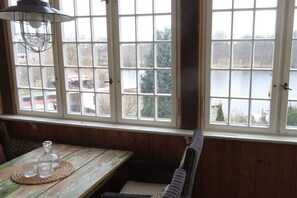 Ein Zimmer mit einem Holztisch, einem Korbstuhl und Blick auf einen See durch große Fenster.