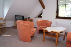 A room with a sloped ceiling, a television, a wooden table, and two orange chairs. - Holiday apartment in Klink with swimming pool (Klink)