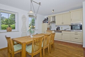 A kitchen with wooden cabinets, a dining table with chairs, a clock, and a hanging light fixture. - Lea semi-detached house (Wiek auf Ruegen)