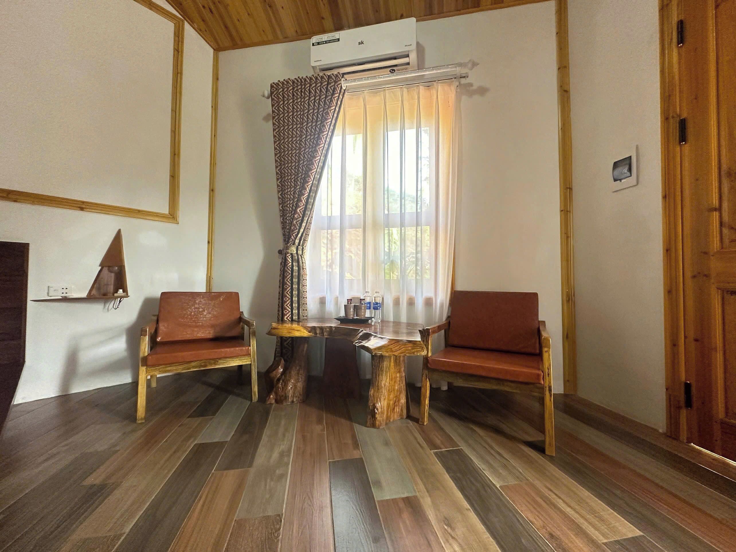 A room with two wooden chairs, a small wooden table, a mirror on the wall, and a window with patterned curtains.
