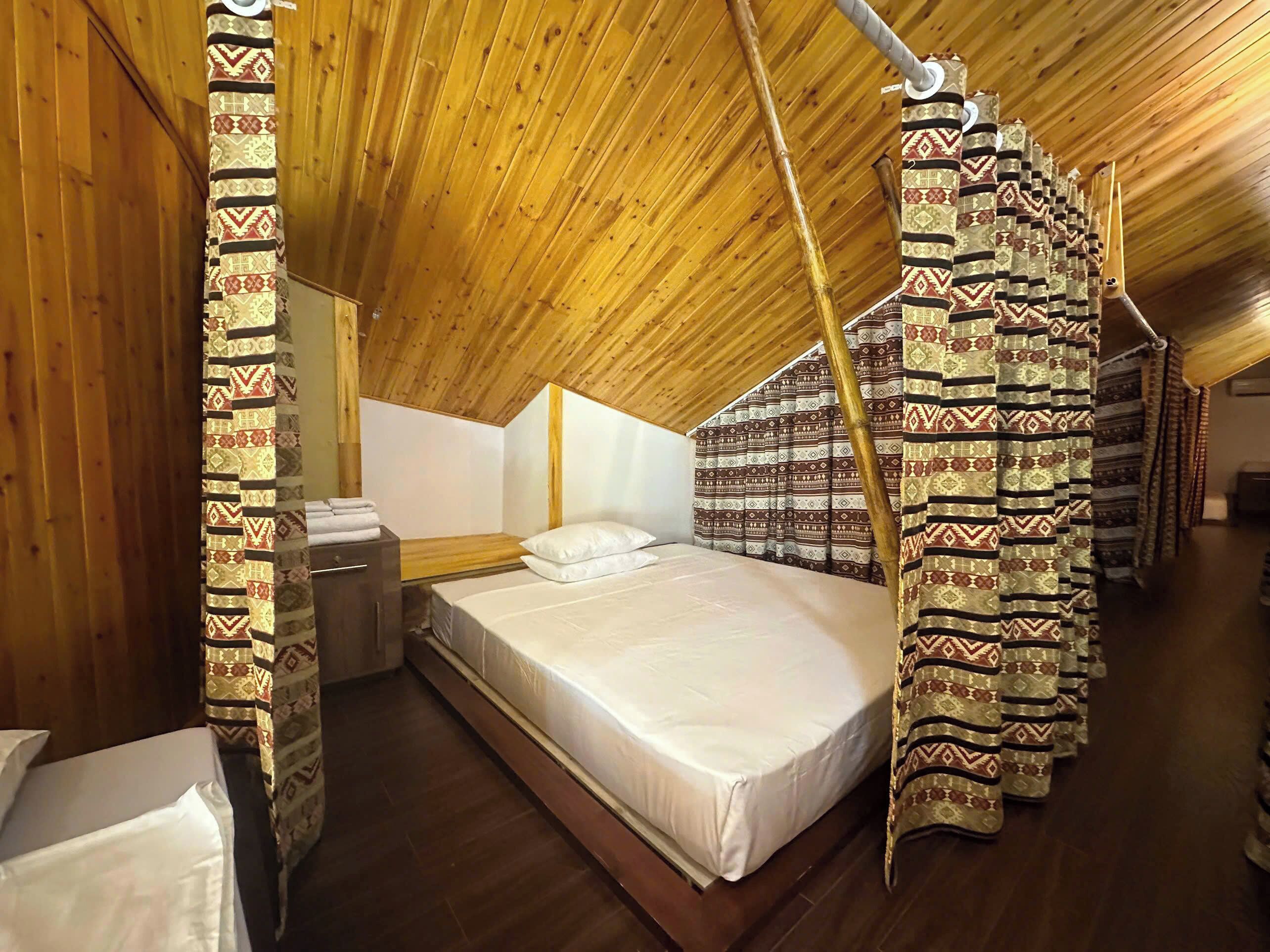 A room with a sloped ceiling, a bed with a white mattress, and patterned curtains.