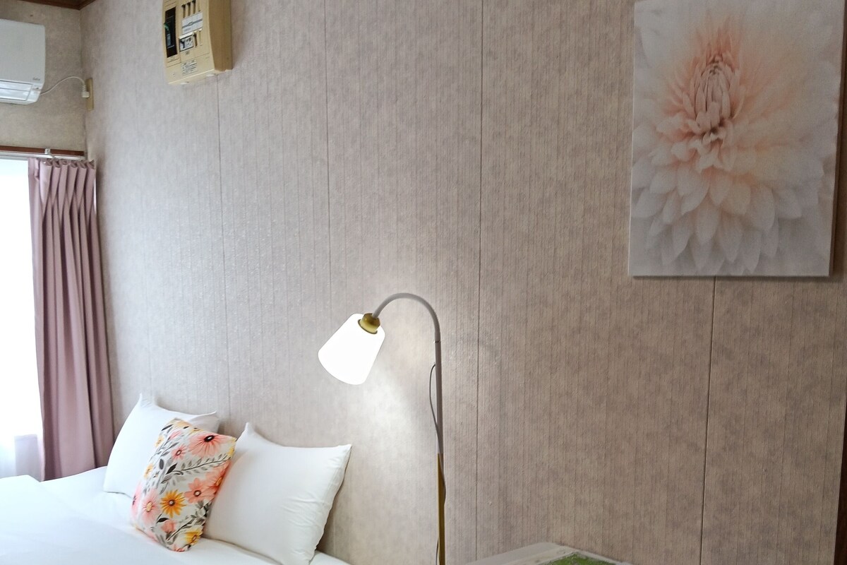Habitación de hotel con cama, escritorio, dos sillas, lámpara, aire acondicionado de pared y una obra de arte floral.