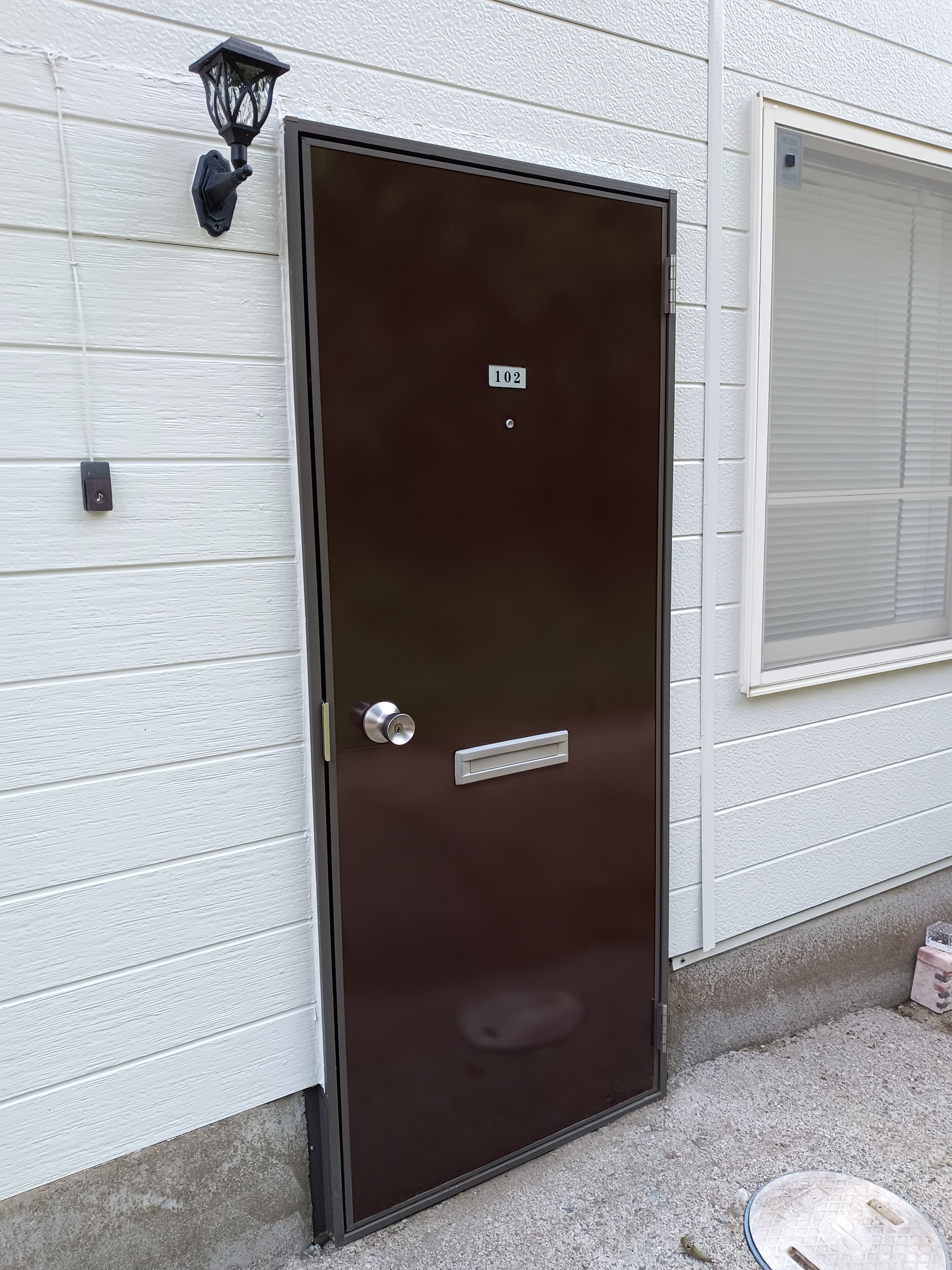 A dark brown door with a number plate, a mail slot, and a wall-mounted light fixture.