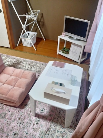 A room with a pink sofa, a small white table with a tissue box and a remote control, a white TV on a stand, and a white laundry basket.