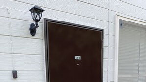 A dark brown door with a number plate, a mail slot, and a wall-mounted light fixture. - guest house mami (Taketa)