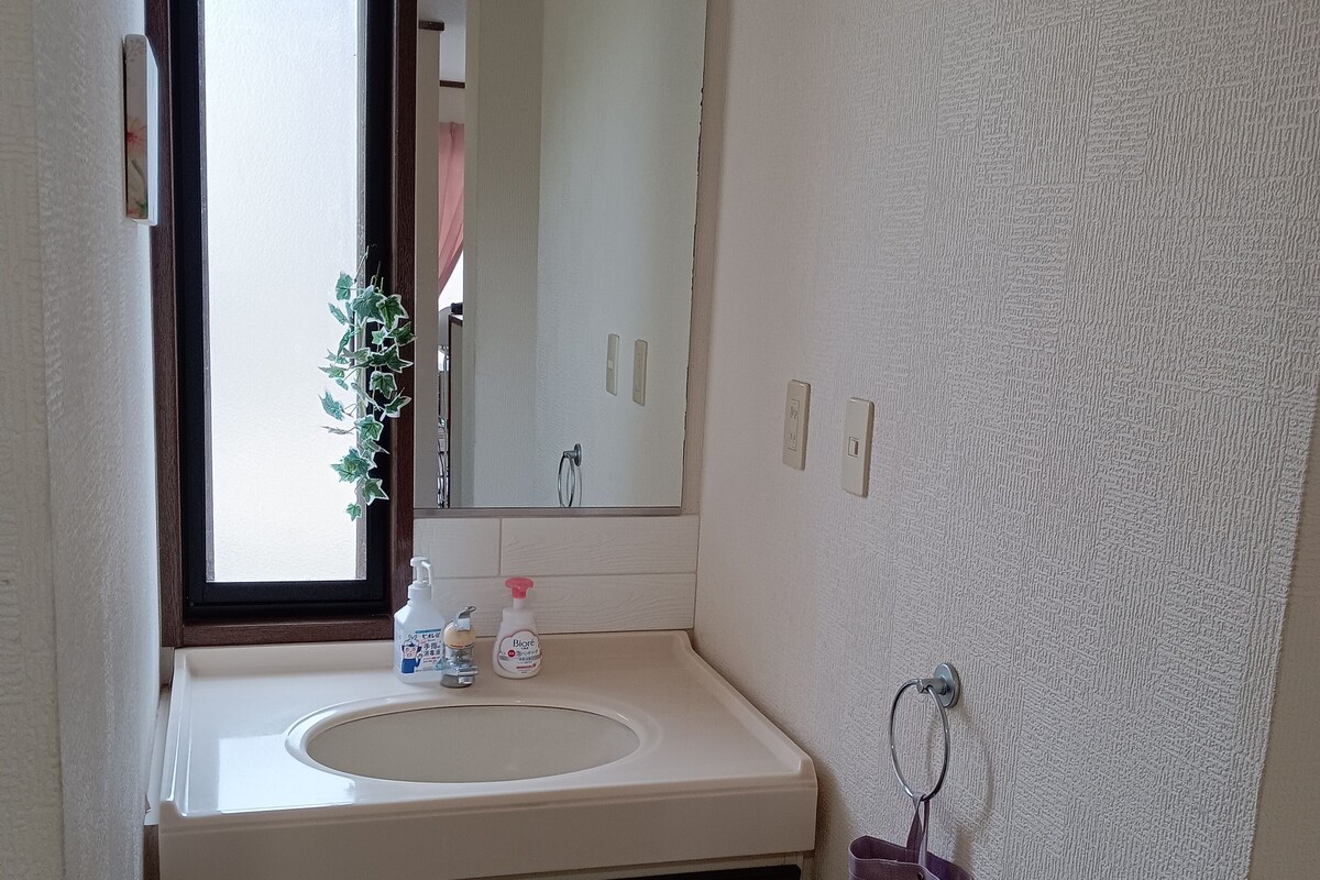 Un baño con un lavamanos blanco, un espejo, una ventana, un perchero para toallas y un cubo de basura.