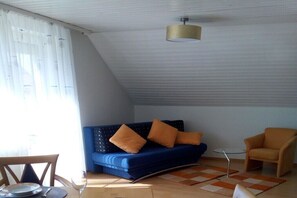 Una habitación con un sofá azul, un sillón naranja, una mesita pequeña y una lámpara en el techo.