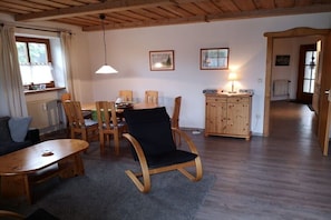 Ein gemütliches Wohnzimmer mit einem Holzesstisch, Stühlen, einem Schaukelstuhl, einem Kamin und einer Küchenzeile mit Holzausbau und einer Pendelleuchte.
