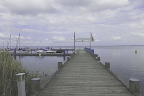 Eine Holzstege führt zu einem Yachthafen mit vertäuten Booten, ein Schild weist auf ein Wassererlebnis-Ziel hin, und ein ruhiger See erstreckt sich unter einem bewölkten Himmel.