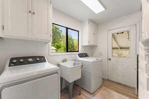 A laundry room with a washing machine, dryer, and a sink. There is a window with a view of trees and a door with an exit sign. - The Hemlock House | The Disconnect Cabin (Dunsmuir)