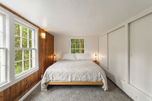 A bedroom with a wooden floor, a bed with a white quilt, two bedside tables with lamps, and a window with a view of trees. - The Hemlock House | The Disconnect Cabin (Dunsmuir)