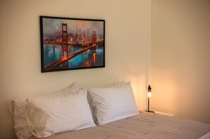 A hotel room with a bed, two white pillows, a nightstand with a lamp, and a framed cityscape painting on the wall. - The Hemlock House | The Disconnect Cabin (Dunsmuir)