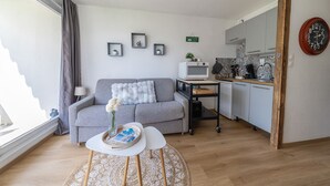 Sala de estar con sofá gris, una mesita redonda con bandeja, una lámpara blanca y una zona de cocina con microondas, lavaplatos y armarios de madera.