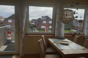 A dining area with a table, chairs, and a view of houses through large windows. - Lighthouse on the sandy beach (Niedersachsen - Norden)