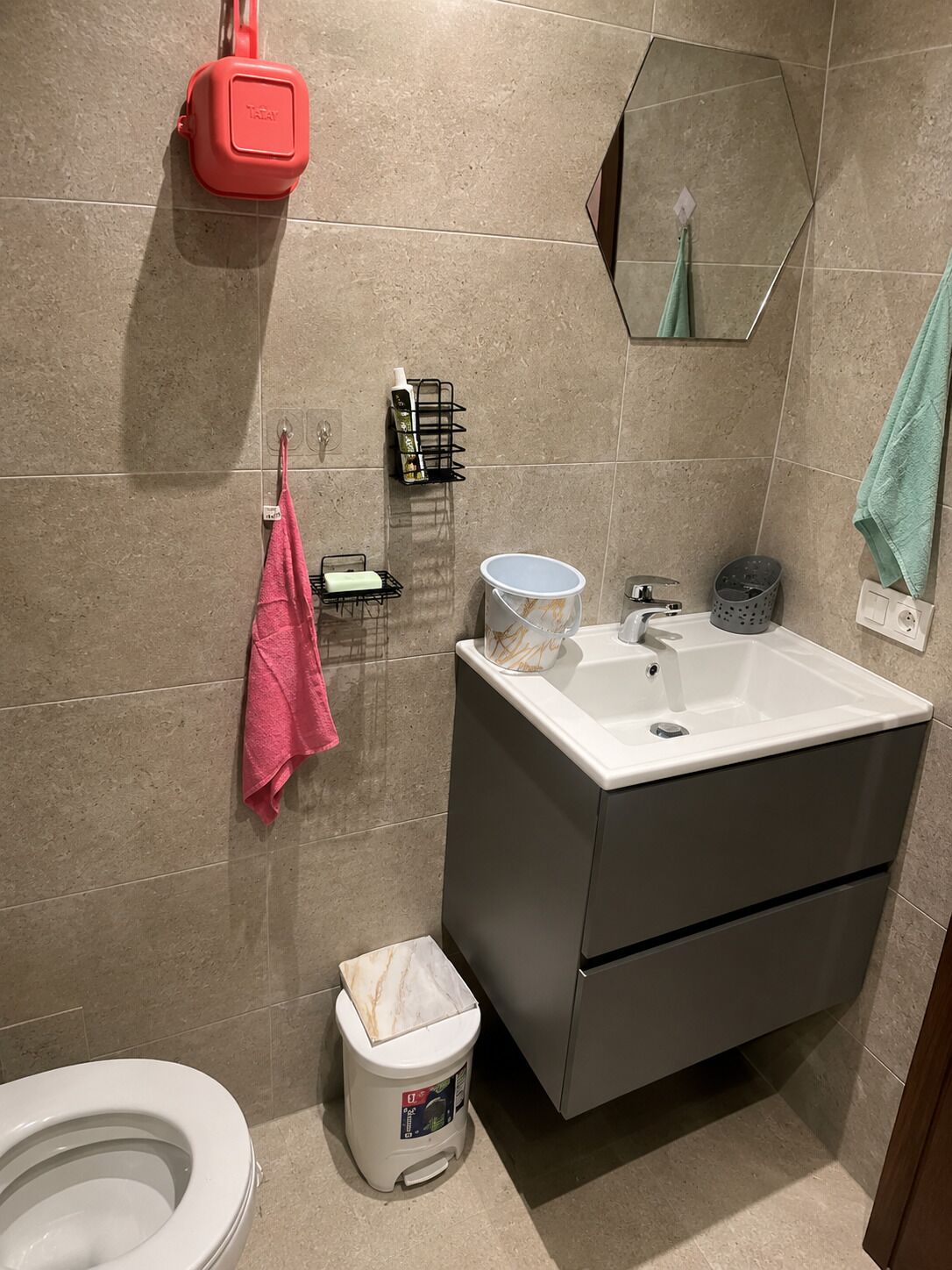 A bathroom with a white sink, a toilet, a trash bin, a towel rack with a pink towel, a soap dispenser, and a mirror.