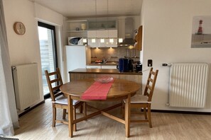 A modern kitchen with white cabinets, a wooden dining table with chairs, a red placemat, and a clock on the wall. - Jade View, Ground Floor (Varel\/Dangast)