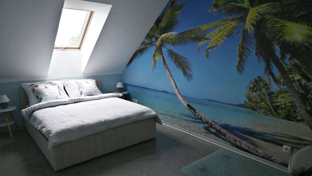 Ein Schlafzimmer mit einem Bett, einem Nachttisch, einer Lampe, einem Fenster und einem Wandgemälde, das eine Strandansicht zeigt.