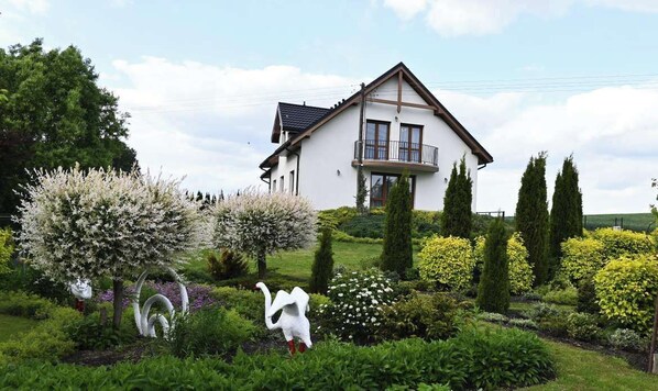 Una casa de dos pisos con balcón, rodeada de un jardín cuidado con diversos arbustos y árboles.