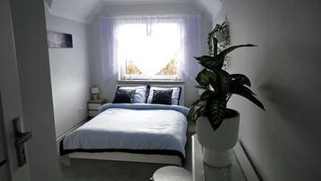 A bedroom with a bed, a bedside table, a nightstand, a window with curtains, and a potted plant.