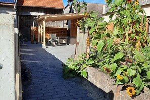 Eine Terrasse mit einer Holz-Pergola, einem Stein-Pflanzgefäß mit Sonnenblumen und einem Ziegelweg, der zu einem Holzzaun führt.