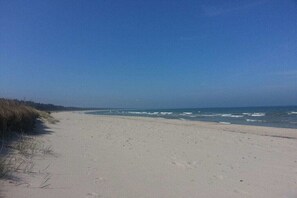 Ein sandiger Strand mit Wellen, blauem Himmel und spärlicher Vegetation.