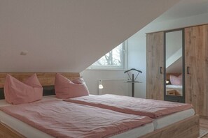 Ein Schlafzimmer mit schrägem Dach, einem Bett mit rosa Bettwäsche, einem Holzschrank und einem Fenster, das natürliches Licht hereinlässt.