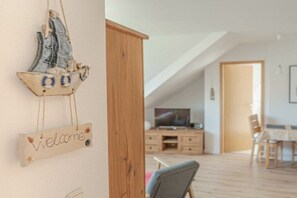 Ein Zimmer mit einem Holzkasten, einem Fernseher auf einem Holzständer, einem Esstisch mit Stühlen und einer Segelboot-Dekoration an der Wand.